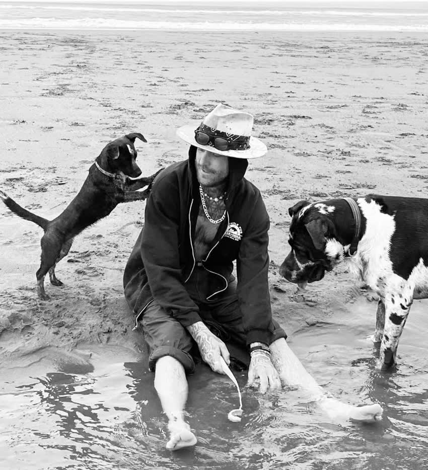 Black and white photo of Scoop sitting in shallow water on a beach in a wide-brim hat, with Bay Bay jumping up at his shoulder and Dude standing beside him