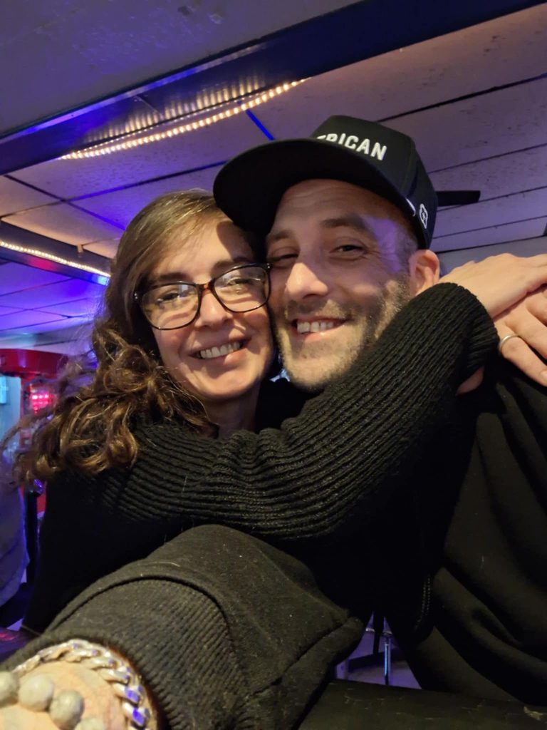 Scoop in a black cap hugging Beth, who has curly brown hair and glasses, both smiling